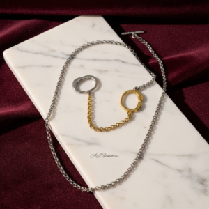 Linked Verse Stainless Steel Gold Silver Necklace with dual-tone chain and interlocking organic rings, displayed on a white marble slab over rich burgundy satin fabric.