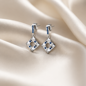 Pair of Square of Secrets 925 silver drop earrings with geometric diamond-shaped pendants and blue sapphire accents, displayed on soft champagne satin fabric.