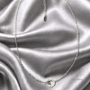 Roma Vitae Stainless Steel Silver Pendant Necklace laid flat on a silver silk background, showing the full length of the silver-toned chain, adjustable extender, and circular Roman numeral pendant with a crystal-studded ring at the center.